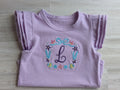 Close up of lavender ruffle tee with embroidered under the sea monogram wreath featuring seashells starfish and bow