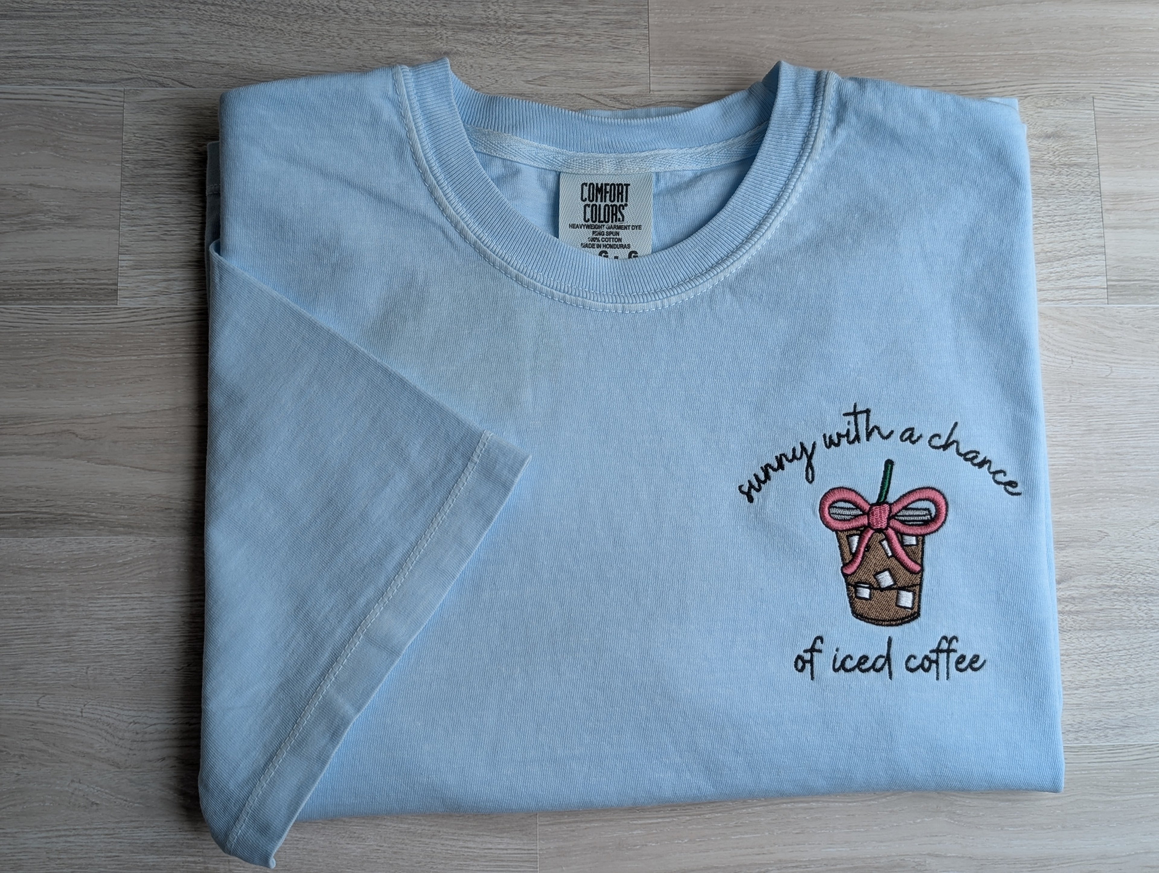 Folded light blue Comfort Colors tee with sunny with a chance of iced coffee embroidered design