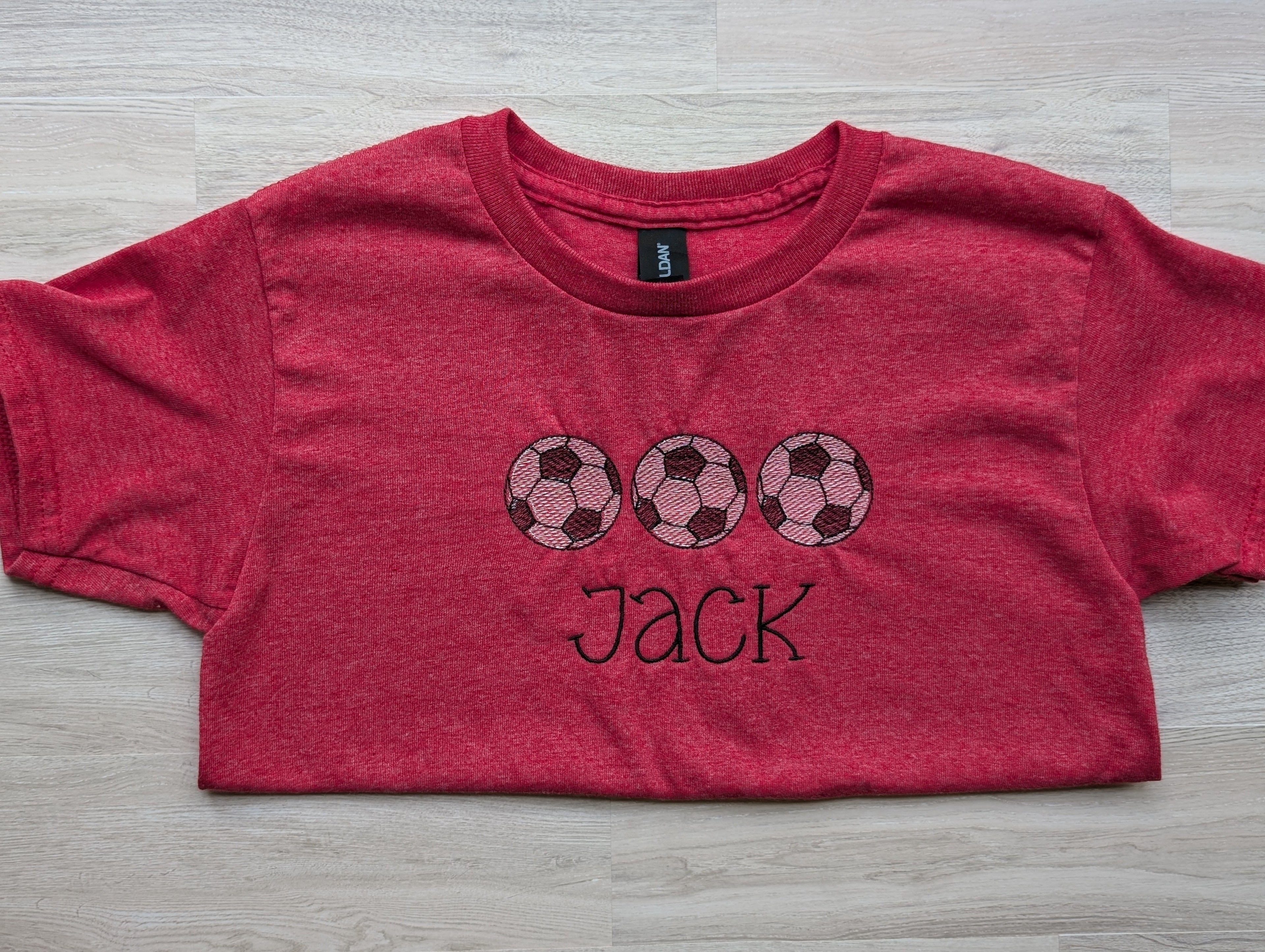 Heathered red kids tee with personalized soccer ball trio embroidery, name Jack, flat lay