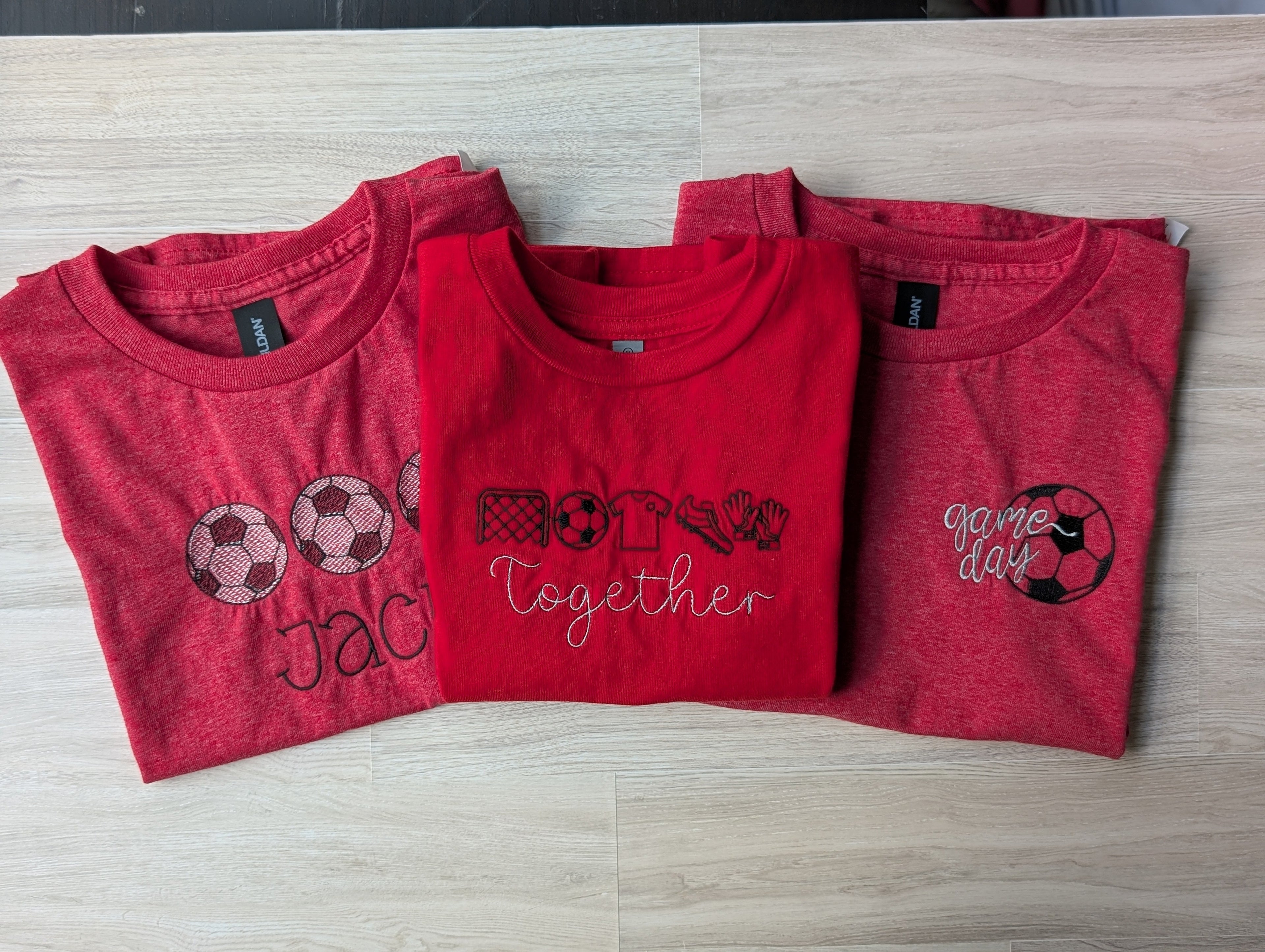 Three red soccer embroidered kids tees stacked showing different designs