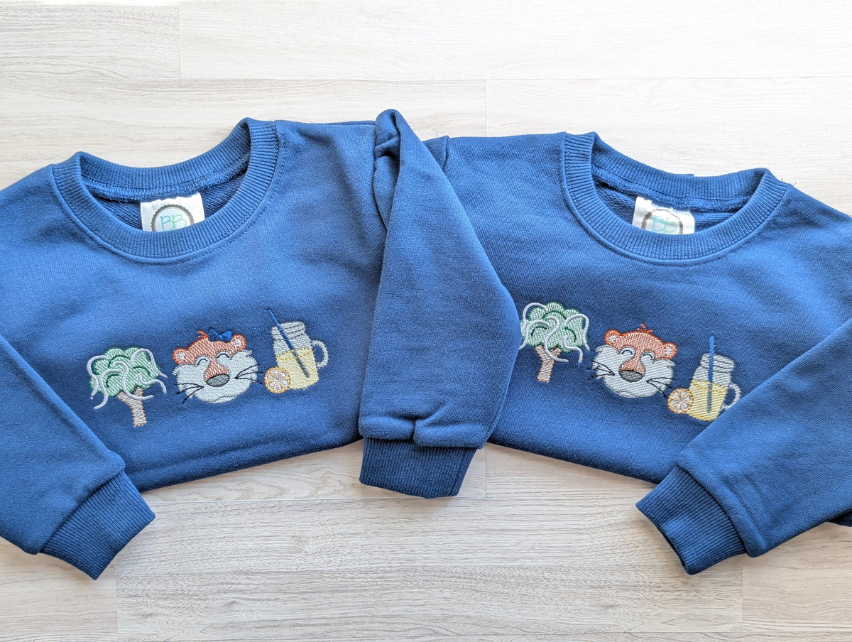 Close view of embroidered oak tree, tiger, and lemonade motifs on a blue sweatshirt