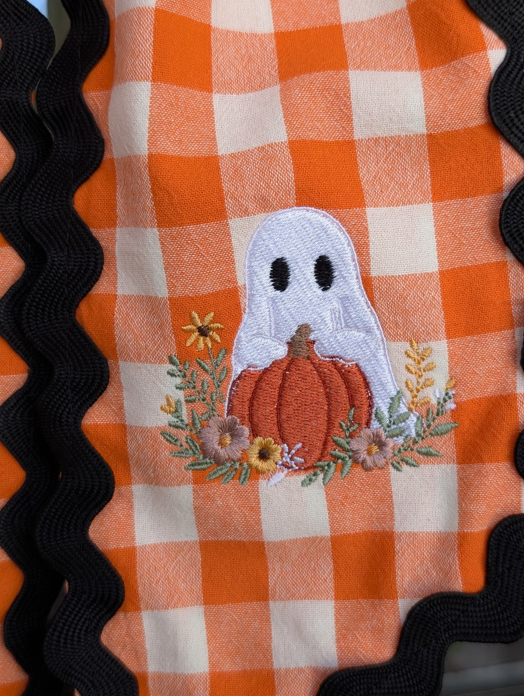 Ghost holding a pumpkin with fall flowers on an orange gingham wreath sash
