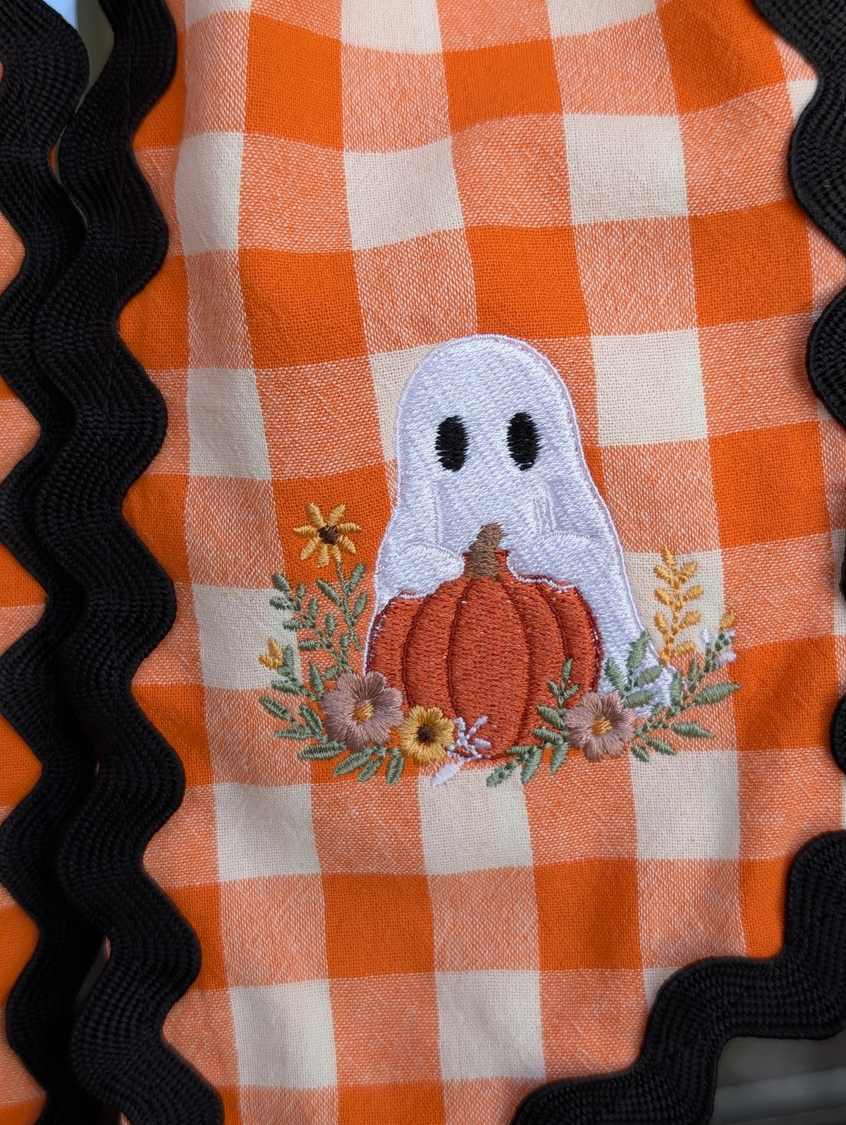Ghost holding a pumpkin with fall flowers on an orange gingham wreath sash